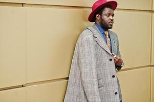 élégant modèle d'homme afro-américain en manteau gris, cravate de veste et chapeau rouge posé sur fond doré. photo