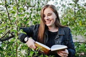 jeune fille brune en jeans contre l'arbre de fleurs de printemps a lu le livre. photo