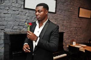 un homme afro-américain puissant et puissant en costume noir renifle une fleur dans un tube à essai contre un piano. photo