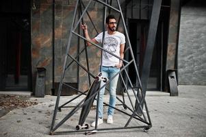 homme arabe de style rue à lunettes avec longboard posé à l'intérieur de la construction pyramidale en métal.