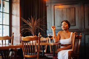 femme afro-américaine, coiffure rétro en robe blanche au restaurant avec verre de vin. photo