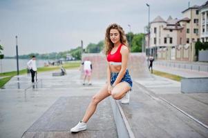 fille modèle bouclée sexy en haut rouge, short en jean jeans, lunettes et baskets posées au skatepark. photo