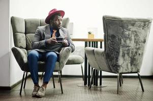 élégant modèle d'homme afro-américain en veste grise cravate et chapeau rouge boire du café au café. photo