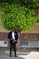 bel homme afro-américain à la mode en tenue de soirée et noeud papillon avec bâton de marche rester sous l'arbre. photo