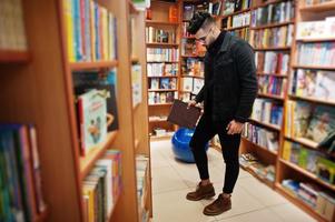 grand étudiant arabe intelligent, portant une veste en jean noir et des lunettes, à la bibliothèque avec un livre à portée de main. photo