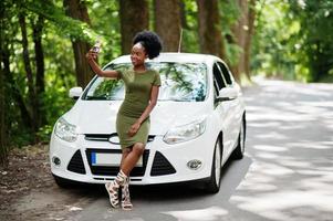 femme afro-américaine posée contre une voiture blanche sur une route forestière et regardant un téléphone portable. photo