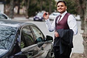 heureux propriétaire de voiture avec des clés à portée de main. homme d'affaires indien élégant en tenue de soirée debout contre une voiture d'affaires noire dans la rue de la ville. photo