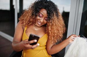 femme afro-américaine glamour en robe jaune et cape de laine blanche assise sur une chaise et regardant sur un téléphone portable. photo