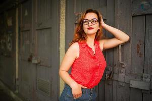 jolie femme rousse à lunettes, porter sur un chemisier rouge et une jupe en jean posant. photo