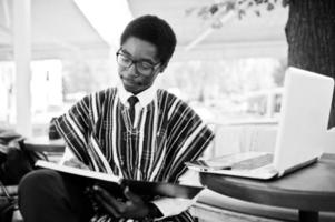 homme africain en vêtements traditionnels et lunettes assis derrière un ordinateur portable au café en plein air et regardant sur son ordinateur portable. photo