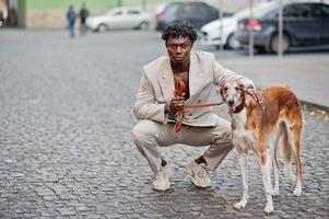 homme afro élégant en costume beige old school avec chien barzoï russe. jeune homme africain à la mode en veste décontractée sur torse nu. photo