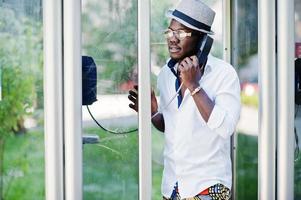 homme afro-américain élégant en chemise blanche et pantalon coloré avec chapeau et lunettes posés en plein air à la cabine téléphonique. garçon modèle à la mode noir. photo