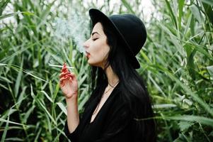 sensuelle fille fumeuse tout en noir, lèvres rouges et chapeau. femme dramatique goth fumant sur roseau commun. photo