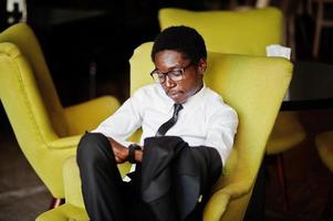 homme d'affaires afro-américain portant une chemise blanche, une cravate et des lunettes au bureau, assis sur une chaise et regardant des montres. photo