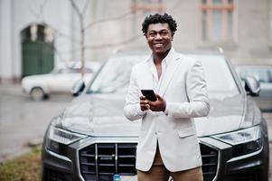 réfléchie jeune beau gentleman afro-américain en tenues de soirée. homme modèle élégant noir en veste blanche avec téléphone portable à mains contre voiture d'affaires. photo