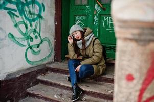 portrait de jeune fille brune en écharpe et chapeau gris, lunettes assis contre l'entrée urbaine. photo