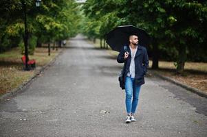 grand homme à barbe arabe à la mode portant un manteau noir avec parapluie et étui à sac posé le jour de la pluie. photo