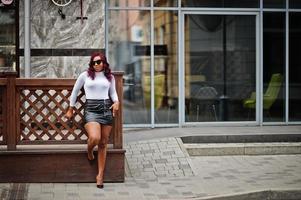 big mama modèle afro-américain taille plus en lunettes de soleil noires et jupe en cuir posée à l'extérieur. photo