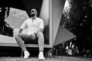 élégant grand modèle d'homme arabe en chemise blanche, jeans et lunettes de soleil assis dans la rue de la ville. Barbe mec arabe attrayant. photo
