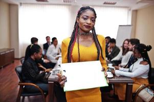 visage de belle femme d'affaires africaine en robe jaune, tenant le presse-papiers sur le fond de la réunion de l'équipe multiraciale des gens d'affaires, assis dans la table de bureau. photo