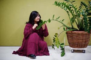 jolie femme sud-asiatique en robe rouge foncé posée au studio sur fond vert avec de la verdure. photo