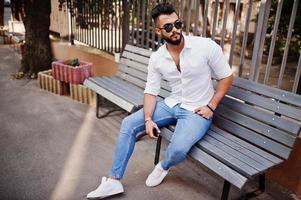 grand modèle d'homme arabe élégant en chemise blanche, jeans et lunettes de soleil posés dans la rue de la ville. Barbe mec arabe attrayant assis sur un banc.