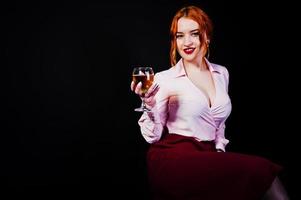 magnifique fille aux cheveux rouges en chemisier rose et jupe rouge avec un verre de vin à portée de main isolé sur noir. photo