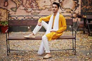 homme élégant indien en vêtements traditionnels jaunes avec foulard blanc posé en plein air sur un banc. photo