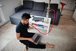 homme indien beau et à la mode en noir assis dans la chambre, fumant du narguilé et travaillant sur un ordinateur portable. photo