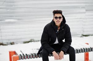 jeune homme indien décontracté en veste noire et lunettes de soleil posées le jour de l'hiver et assis sur un banc. photo