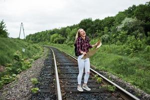 portrait d'une jolie fille blonde en chemise tartan marchant sur le chemin de fer avec une carte dans les mains. photo