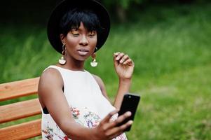 incroyable femme modèle afro-américaine en pantalon vert et chapeau noir posé sur un banc au parc et faisant selfie sur téléphone mobile. photo