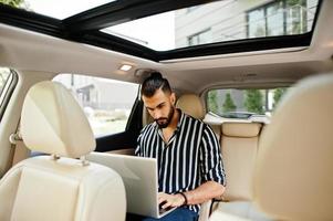 un homme arabe réussi porte une chemise rayée et des lunettes de soleil pose à l'intérieur d'une voiture suv blanche avec un ordinateur portable dans les mains. photo