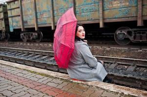 fille brune en manteau gris avec parapluie rouge dans la gare. photo