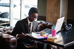 homme d'affaires afro-américain portant un costume noir et des lunettes assis au bureau avec un ordinateur portable et travaillant. photo