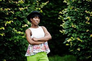 incroyable femme modèle afro-américaine en pantalon vert et chapeau noir posé au parc. photo