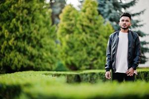 homme barbe indien élégant à la veste en cuir noir. modèle indien posé au jardin verdoyant de la ville. photo