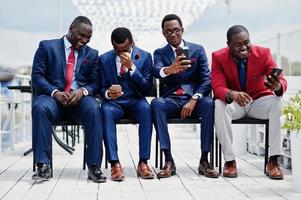 groupe de quatre hommes afro-américains heureux et réussis en costume. riches hommes d'affaires noirs assis sur des chaises en regardant des téléphones portables. photo