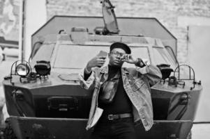 homme afro-américain en veste de jeans, béret et lunettes, avec un cigare posé contre un véhicule blindé militaire btr, et faisant un selfie sur un téléphone portable. photo