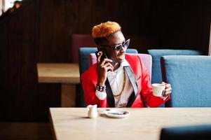 modèle d'homme afro-américain de mode en costume rouge, avec des cheveux en surbrillance et des lunettes de soleil assis au café avec du café et parlant au téléphone mobile. photo