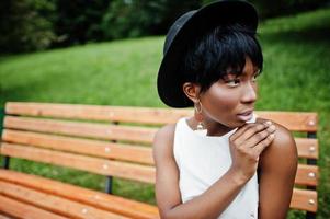 incroyable femme modèle afro-américaine en pantalon vert et chapeau noir posé sur un banc au parc. photo