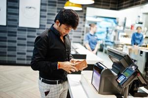 homme indien tenant un portefeuille derrière sa caisse avec écran de commande et terminal de paiement par carte dans un café alimentaire. photo