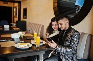 charmant couple indien amoureux, porter un sari et un costume élégant, assis au restaurant et regardant le téléphone ensemble. sur les gâteaux de dessert de table, la crème glacée et le jus. photo