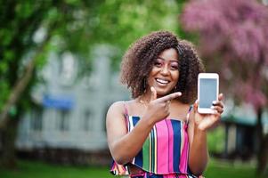 femme afro-américaine à la mode en combinaison rayée rose posée dans la rue de la floraison printanière et montre le doigt à l'écran du téléphone portable. photo