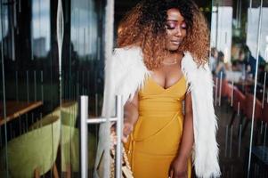 femme afro-américaine glamour en robe jaune et cape en laine blanche avec sac à main posé à l'entrée de la porte vitrée du restaurant. photo