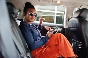 riche femme africaine d'affaires sur des lunettes de soleil s'asseoir à la voiture suv avec des sièges en cuir noir. téléphone portable à portée de main. photo