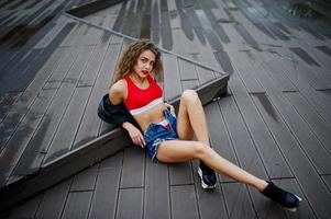 fille modèle bouclée sexy en haut rouge, short en jean, veste en cuir et baskets posées sur le sol. photo
