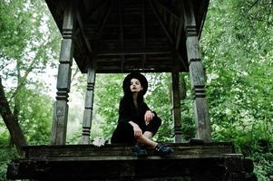fille sensuelle tout en noir, lèvres rouges et chapeau. femme dramatique goth assise sur une arche en bois à la forêt. photo