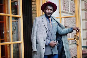 élégant modèle d'homme afro-américain en manteau gris, cravate de veste et chapeau rouge contre la porte d'entrée du café. photo