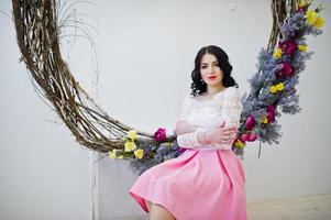 jeune fille brune en jupe rose et chemisier blanc posé à l'intérieur contre une grande couronne décorée. photo